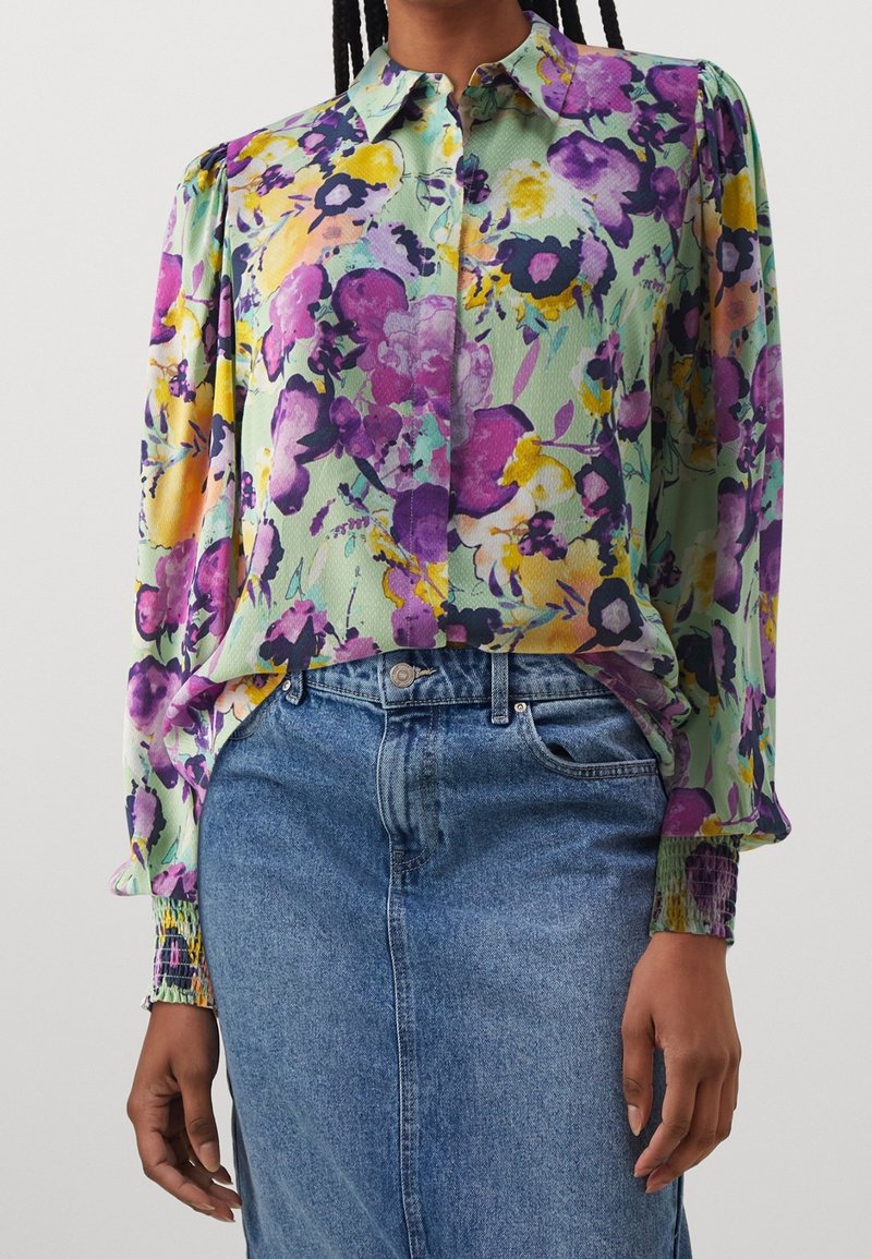 Femme portant un chemisier fleuri boutonné avec des fleurs jaunes, violettes et roses, associé à une jupe en jean bleue taille haute.