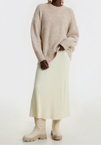 Maglione beige a coste con un collo alto, abbinato a una gonna midi plissettata crema e stivaletti beige con suole testurizzate.