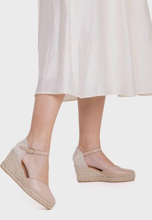 Mujer que lleva alpargatas de cuña beige con tiras en el tobillo y una falda midi blanca, de pie sobre un fondo blanco.