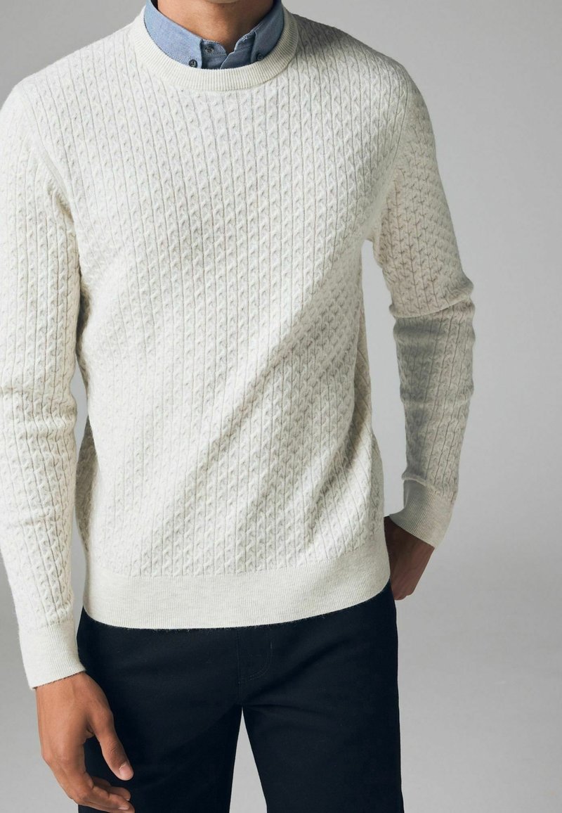 Cremefarbener Strickpullover mit strukturiertem Diamantmuster, Rundhalsausschnitt und gerippten Bündchen. Über einem hellblauen Hemd mit Kragen getragen.