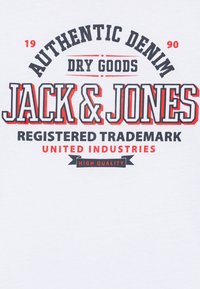 Bjela majica s podebljanim crnim i crvenim tipografijom. Tekst uključuje "AUTENTIČNI DENIM", "JACK & JONES", "REGISTRIRANA TRGOVINSKA MARKA" i "VISOKA KVALITETA."