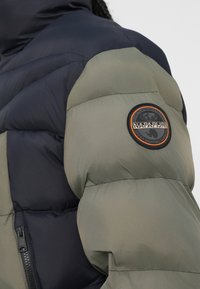 Pufjakke med en marineblå overdel og en olivengrøn underdel. Har et cirkulært logo-patch på ærmet og en lynlåslomme.