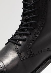 Bota de cuero negro con cordones, que presenta una textura suave, punta reforzada y sutiles detalles de costura. Los cordones son negros con puntas redondeadas.