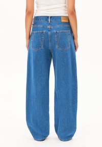Model's back wearing baggy blue jeans with a high waist and large pockets, set against a plain white background.
