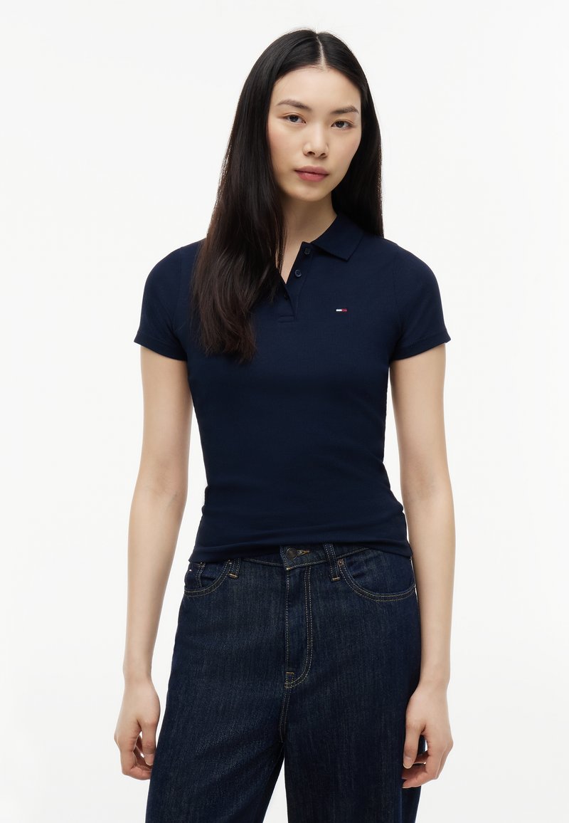 Jeune femme aux longs cheveux foncés portant un polo bleu marine ajusté et un jean bleu foncé, debout devant un fond blanc uni.