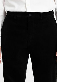 Črne kord stojnike hlače z navpičnimi rebri; imajo gumbasto zapiranje in pasne zanke. Tkanina ima mehak otip in strukturirano obliko.