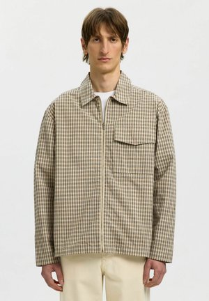 Mladý muž v béžovej kárované bunde na zips nad bielou košeľou a krémovými nohavicami, stojaci proti jednoliatemu bielemu pozadiu.