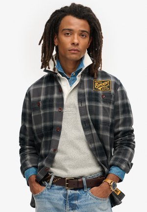 Camicia flanella a quadri nera e grigia, con bottoni rossi e una toppa con logo. Indossata sopra un maglione leggero e una camicia di jeans, abbinata a dei jeans.