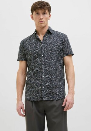 Chemise à manches courtes en tissu sombre avec un motif floral blanc et rouge, patte de boutonnage à l'avant et col classique, portée avec un pantalon gris.
