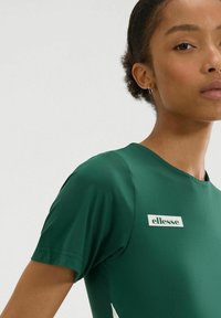 Zielony krótki rękaw bluzki wykonany z gładkiego materiału z prostokątną białą naszywką z logo na piersi. Krój dopasowany.