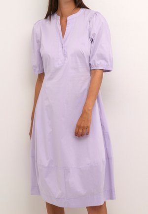Kvinde iført en lys lilla midi-kjole med korte pufærmer og V-udskæring med knaplukning, der står op ad en hvid væg.