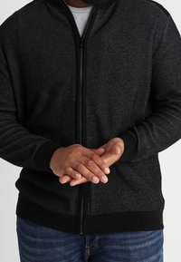Giacca nera con zip, caratterizzata da un design di maglia testurizzata, con collo alto e polsini a costine. Le mani sono incrociate davanti al torso.