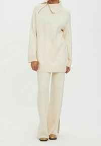 Femme portant une tunique crème à manches longues avec un col montant et un pantalon large crème, associée à des chaussures carrées beige clair.