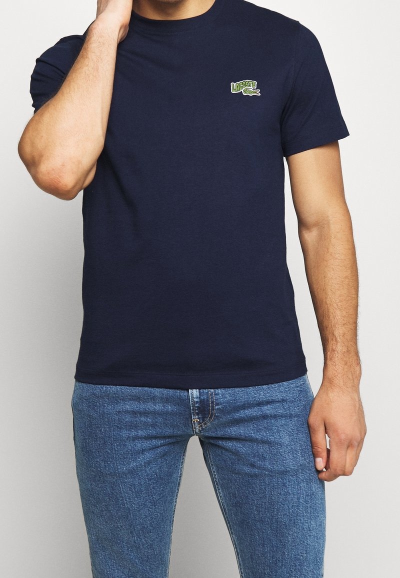 Marinblå bomullströja med rund halsringning, korta ärmar och en grön broderad logotyp på bröstet. Matchad med blå denimjeans.