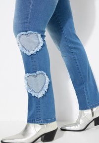 Jeans de mezclilla en azul con parches en forma de corazón desgastados y con bordes deshilachados. Acompañados de botas plateadas metálicas de punta afilada.
