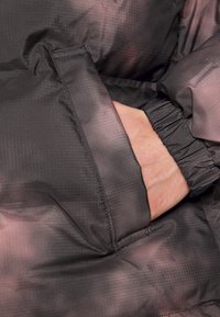 Mano dentro del bolsillo delantero de una chaqueta oscura y estampada, con el puño elástico visible.