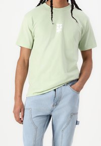 Light green cotton t-shirt with short sleeves and a round neckline, featuring a white logo design on the front. Worn with light blue denim jeans.