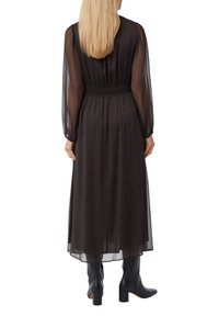 Schwarzes langärmliges Kleid mit geraffter Taille, hergestellt aus transparenter Stoff. Es hat eine fließende Form und wird mit knielangen schwarzen Stiefeln getragen.
