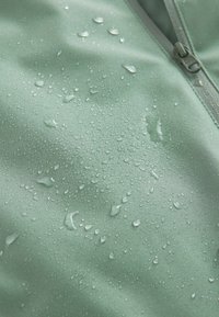 Vesitiivis vihreä kangas, jossa on sileä pinta, ja pinnalla on vesipisaroita sekä vetoketju yhdellä reunalla.