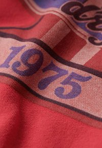 Tessuto rosso con un design caratterizzato da grandi numeri viola "1975" e testo curvo. La texture sembra morbida con un motivo sottile.