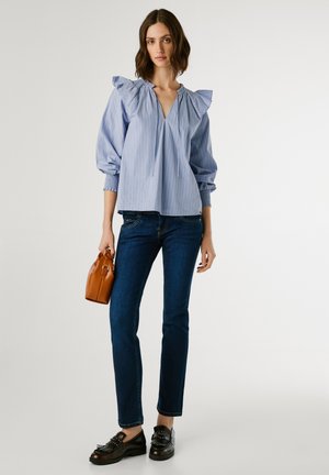 Stribet blå bluse med flæset halsudskæring, lange puffede ærmer, parret med mørke jeans og sorte nitter sko. Holder en lille brun håndtaske.