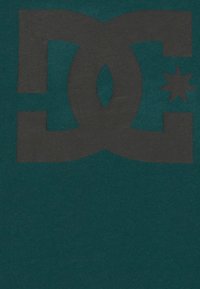 Tyrkysová látka se velkým černým logem DC a akcentem hvězdy. Design je odvážný s matnou texturou a minimalistickým stylem.