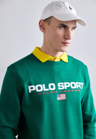 Sweat-shirt vert avec le texte "POLO SPORT RALPH LAUREN" en blanc et rouge, superposé sur un col polo jaune, porté avec une casquette blanche.