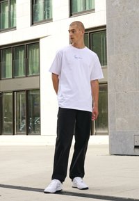 White cotton t-shirt with blue graphic, paired with black trousers and white sneakers, set against a modern urban backdrop.