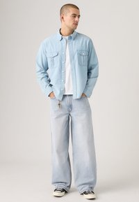 Light blue denim shirt with two chest pockets, unbuttoned at the top. Paired with loose-fitting light blue jeans and black sneakers.