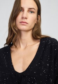 Giovane donna con lungo capelli castani che indossa un cardigan V-neck nero ricoperto di paillettes sopra un top abbinato, su uno sfondo chiaro e semplice.
