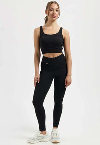 Frau steht vor weißem Hintergrund, trägt ein schwarzes bauchfreies Tanktop, schwarze hoch taillierte Leggings und weiße Sneakers.