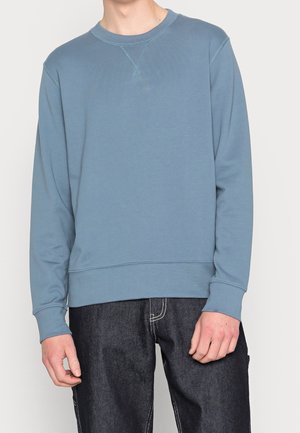 Mann trägt hellblaues Sweatshirt mit Rundhalsausschnitt und dunkelblaue Jeans, steht vor hellem einfarbigem Hintergrund.