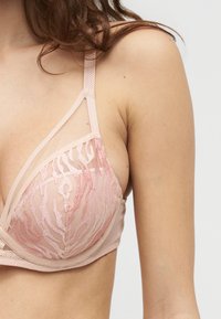 Γυναίκα που φοράει ένα ανοιχτό ροζ διάφανο σουτιέν με κεντητά αφηρημένα μοτίβα και ρυθμιζόμενους ώμους, με απλό φόντο.