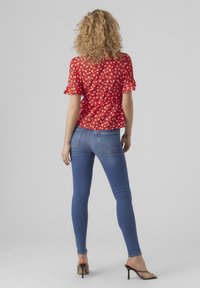 Blusa floreale rossa con maniche corte a volant, abbinata a jeans skinny blu. Il modello è in piedi con le spalle rivolte verso la macchina fotografica, mostrando il design e la vestibilità.