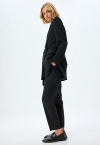 Zwarte op maat gemaakte blazer met een riem, gecombineerd met aangesloten zwarte broek en glanzende zwarte instappers. Glad materiaal, gestructureerd ontwerp en strakke lijnen.
