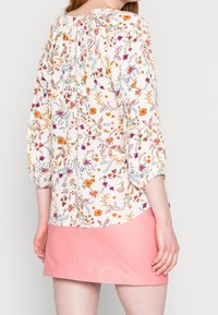 Bluse mit Blumenmuster und puffartigen Ärmeln, auf einem hellen Hintergrund mit bunten Blumenmustern. Kombiniert mit einem rosa Leder-Minirock.