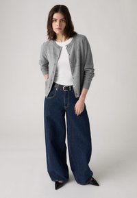 Cardigan en tricot gris avec fermeture à boutons, chemise blanche ajustée, jeans en denim foncé à jambes larges, escarpins noirs à bout pointu, texture lisse, style décontracté.