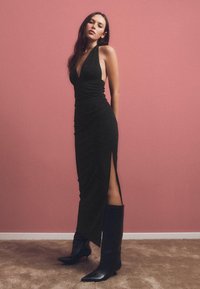 Fekete nyakpántos ruha, testhez álló sziluettel, textúrált anyagból, oldalsó hasítékkal, térdig érő fekete csizmával párosítva, rózsaszín háttér előtt.