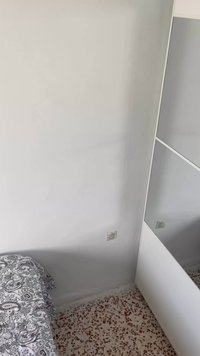 Bijela zidna površina s kutnim dijelom koji sadrži ogledalo i ormarić. Krevet s pokrivačem u crno-bijelom paisley uzorku i pločice na podu.