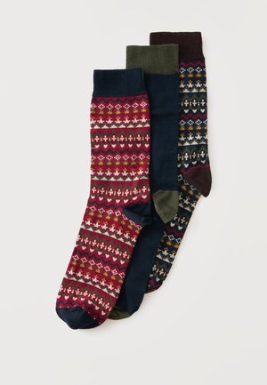 Trois paires de chaussettes à motifs rouges, bleu marine et marron foncé avec des dessins géométriques et floraux, posées en chevauchement sur un fond blanc.