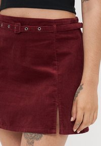 Bordeaux corduroy mini-rok met een vooropening, riemlussen en metalen oogjes. Textiel met structuur en een nette zoomafwerking.