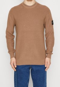 Brązowy sweter o wrabianym wzorze z długimi rękawami i okrągłym dekoltem. Posiada czarną naszywkę z logo na lewym rękawie. Noszony z niebieskimi dżinsami.