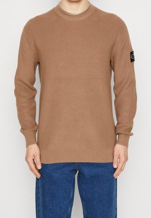 Brun ribbstickad tröja med långa ärmar och rundad halsringning. Inkluderar en svart logotypapplikation på vänster ärm. Bärs med blå jeans.