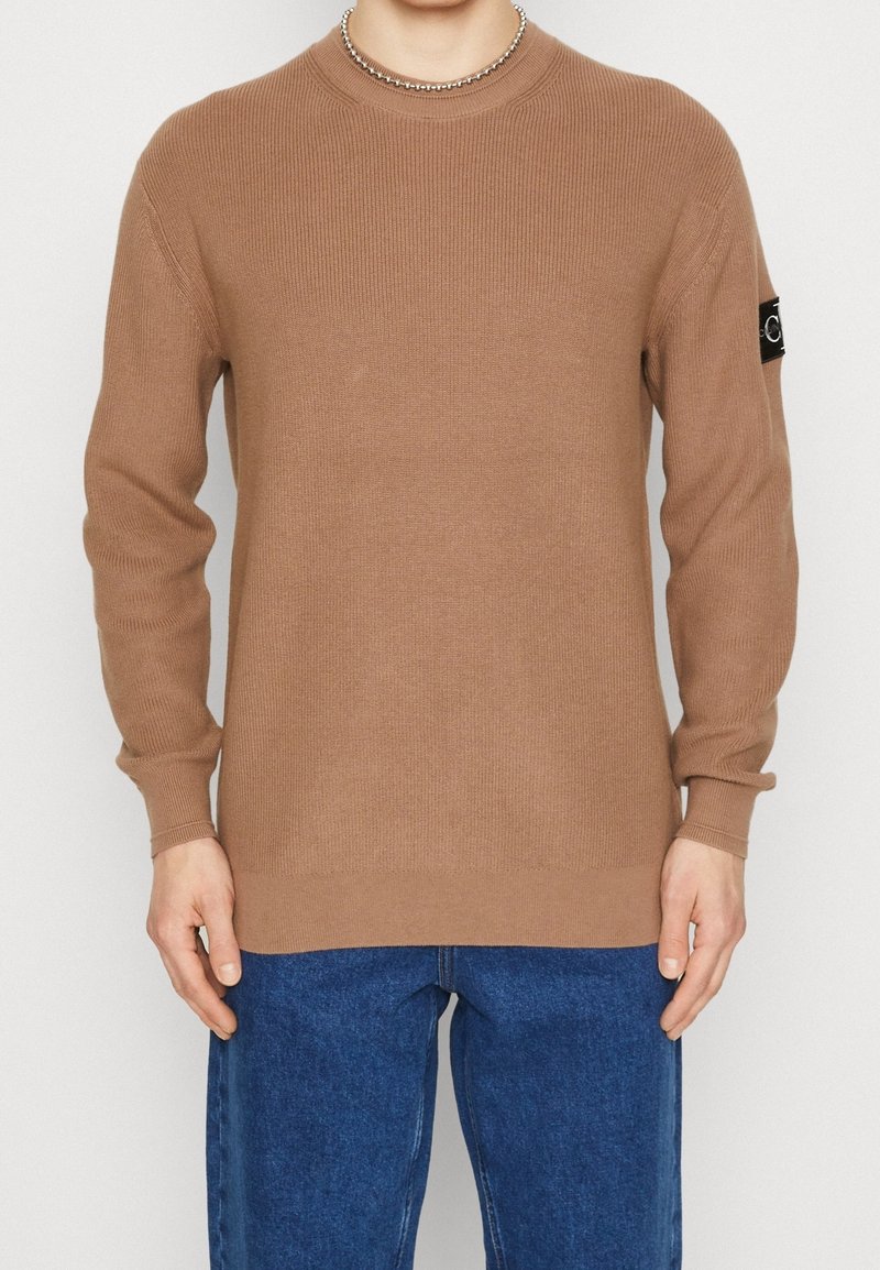 Brązowy sweter o wrabianym wzorze z długimi rękawami i okrągłym dekoltem. Posiada czarną naszywkę z logo na lewym rękawie. Noszony z niebieskimi dżinsami.