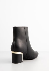 Bottine en cuir noir avec un bout arrondi et un talon bloc orné d'accents dorés. Texture lisse et design épuré, avec une couture latérale sans coutures.