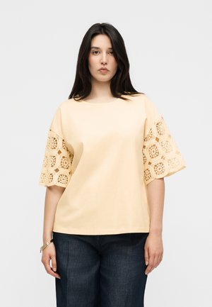 Kvinde med langt mørkt hår iført en bleg gul top med hæklet blondeærmer og mørkeblå jeans mod en ensfarvet grå baggrund.