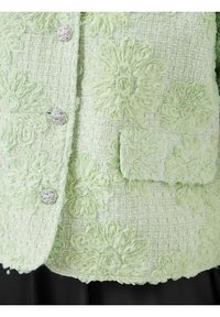 Lys grønn teksturert blazer med blomsterapplikasjoner og sølvknapper. Har en lomme med lignende blomsterdetaljer.