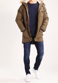 Man som bär en olivgrön huvmantel, mörkblå tröja, slim-fit jeans och vita sneakers, stående mot vit bakgrund.