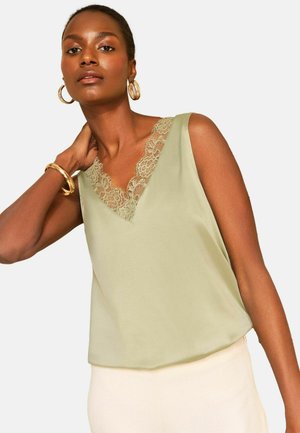 Femme portant un top sans manches vert clair bordé de dentelle, avec des boucles d’oreilles créoles dorées et un bracelet, posant avec la main sur le cou.