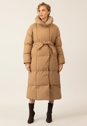 Long, padded coat in a light brown color with a high collar, belted waist, and ribbed cuffs. Features large quilted panels.